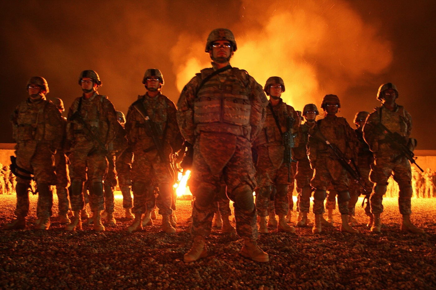 A formation of U.S. Army soldiers in combat gear standing at night in front of a large fire